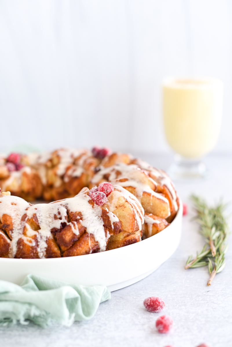Monkey Bread in a ring with eggnog frosting drizzled over the top surrounded by sugared cranberries, rosemary and eggnog.