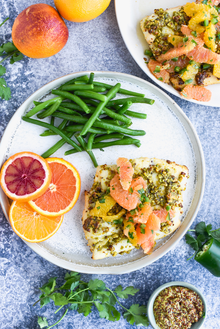 fish topped with oranges, pistachios and herbs on a plate with green beans and orange slices
