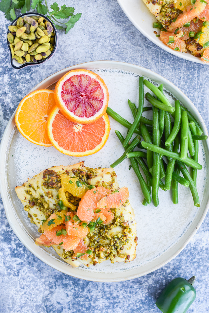 fish topped with oranges, pistachios and herbs on a plate with green beans and orange slices