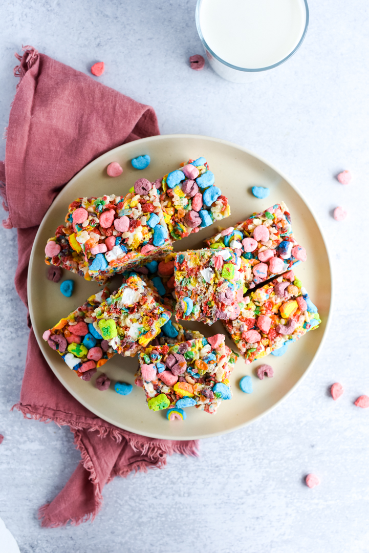 rainbow rice krispie treats stacked on a plate with marshmallows with a glass of milk in the background